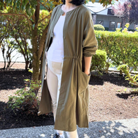 Person wearing a long moss green tunic coat over a white shirt and beige pants, standing outdoors.