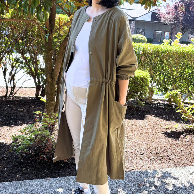 Person wearing a long moss green tunic coat over a white shirt and beige pants, standing outdoors.