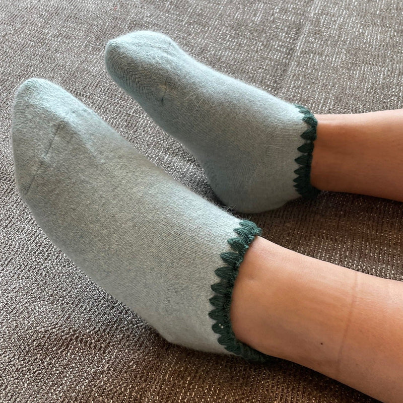 Angora Slipper Socks with anti-slip soles featuring transparent grip dots for extra safety. Each pair is handcrafted, incorporating traditional craftsmanship, and finished with cherrystone shell-shaped embroidery on the sock opening. Soft, cozy, and comfortable for ultimate foot pampering.