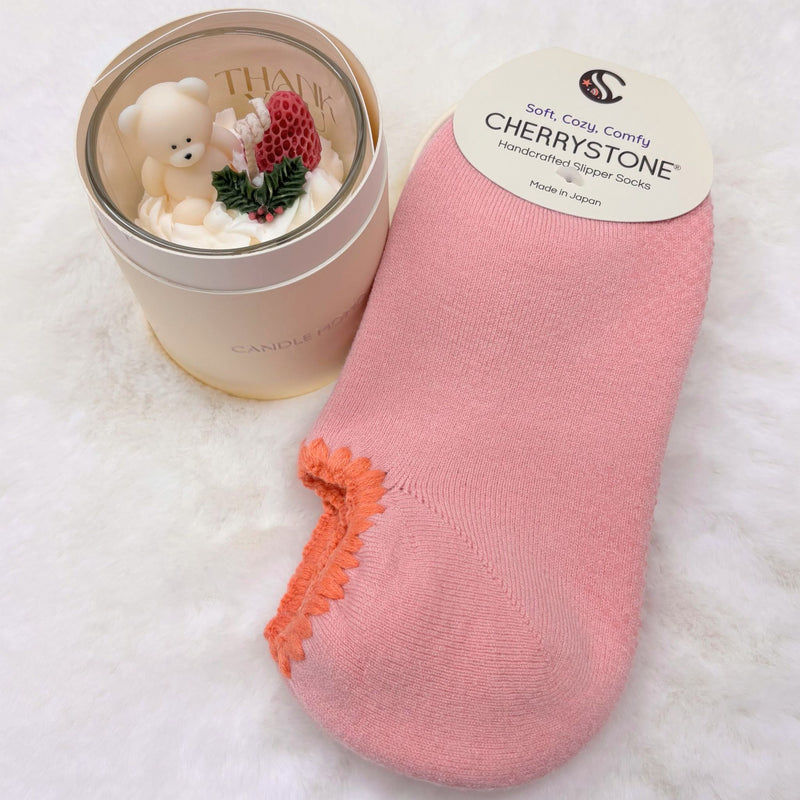 Pink slipper socks with orange trim next to a small candle in a decorative container on a white surface.