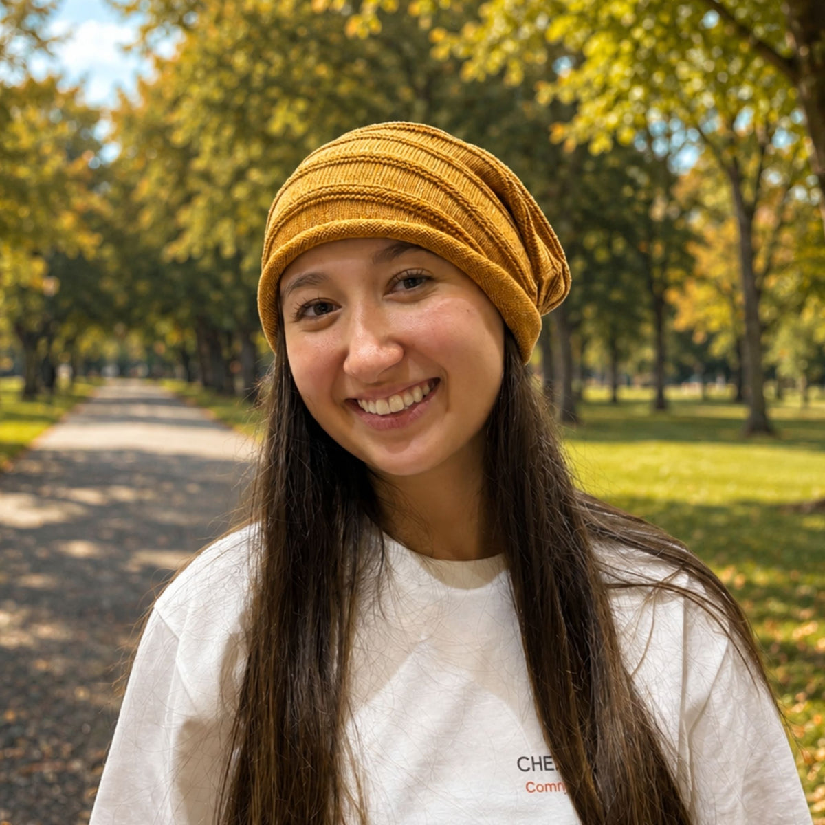 A smiling woman outdoors on a tree-lined path wearing a mustard yellow ribbed cotton & silk blend knit beanie and a white T-shirt.