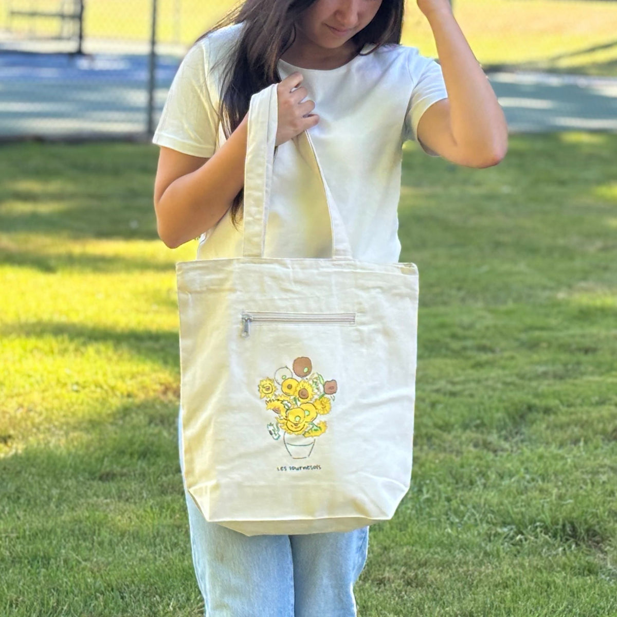 Person holding a white tote bag with a sunflower design outdoors.
