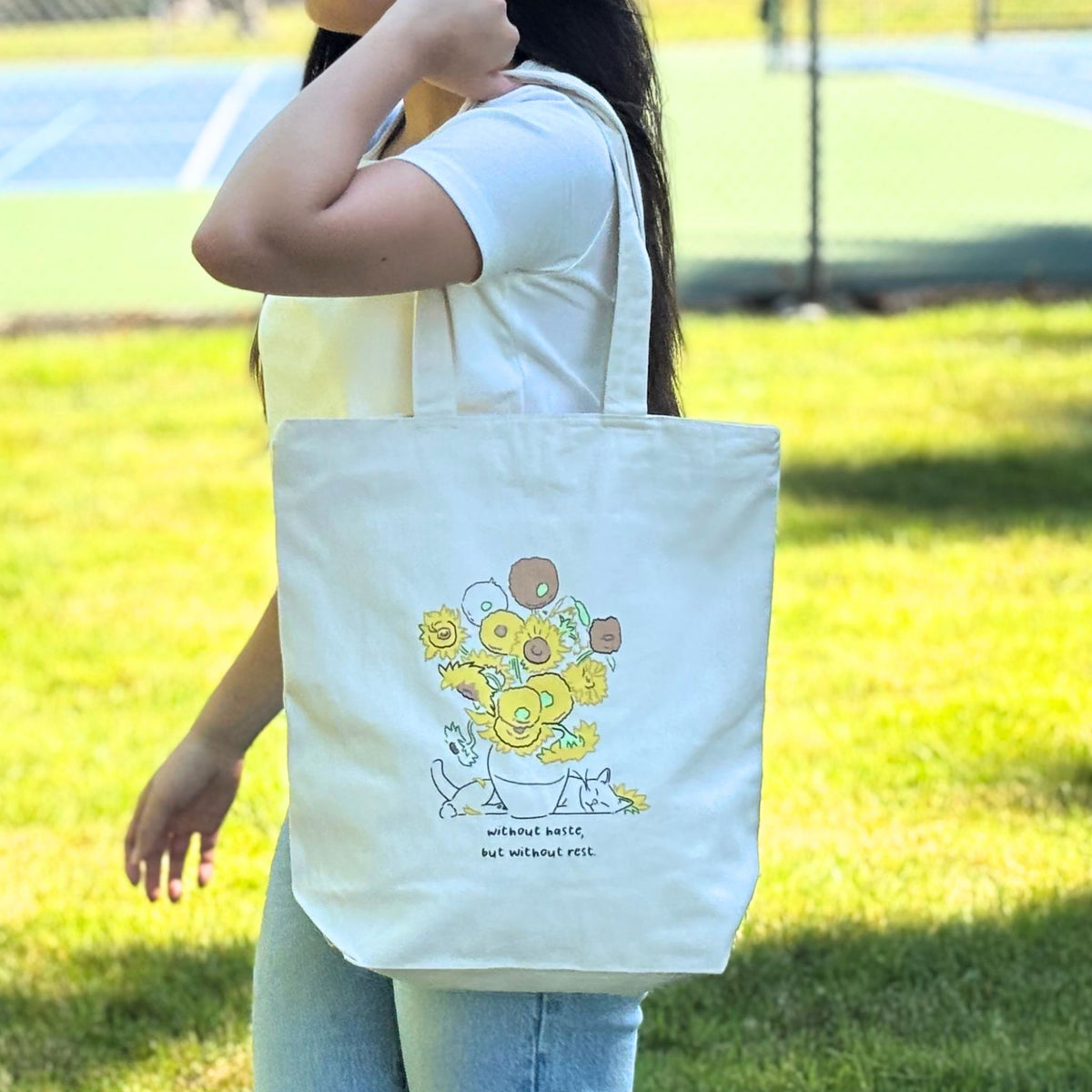 Person holding a white tote bag with a sunflower design outdoors.