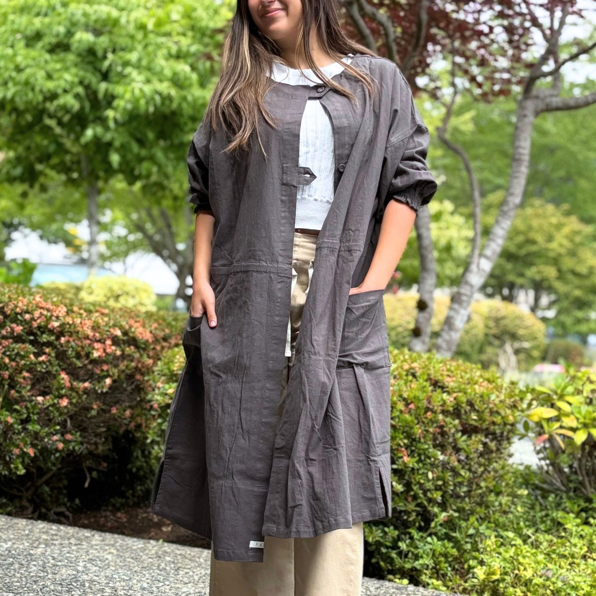 A woman wearing a long charcoal gray coat in an outdoor setting with greenery.