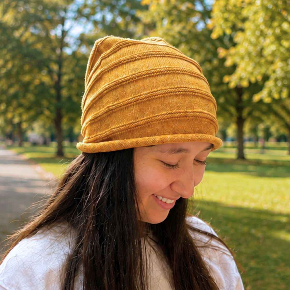 A woman wearing a mustard yellow ribbed cotton & silk blend knit beanie hat, photographed outdoors in a sunny park with blurred green trees in the background.