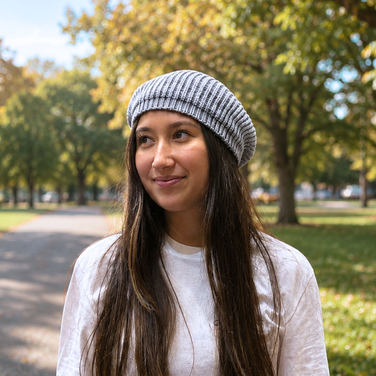 A woman with long dark hair wearing a gray and white striped silk knit beanie and a white t-shirt, standing in a sunny park with trees in the background.