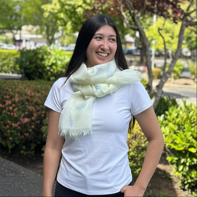 A woman smiling outdoors in front of a red-leafed tree and green bushes, wearing a white short-sleeve top and a light yellow scarf.