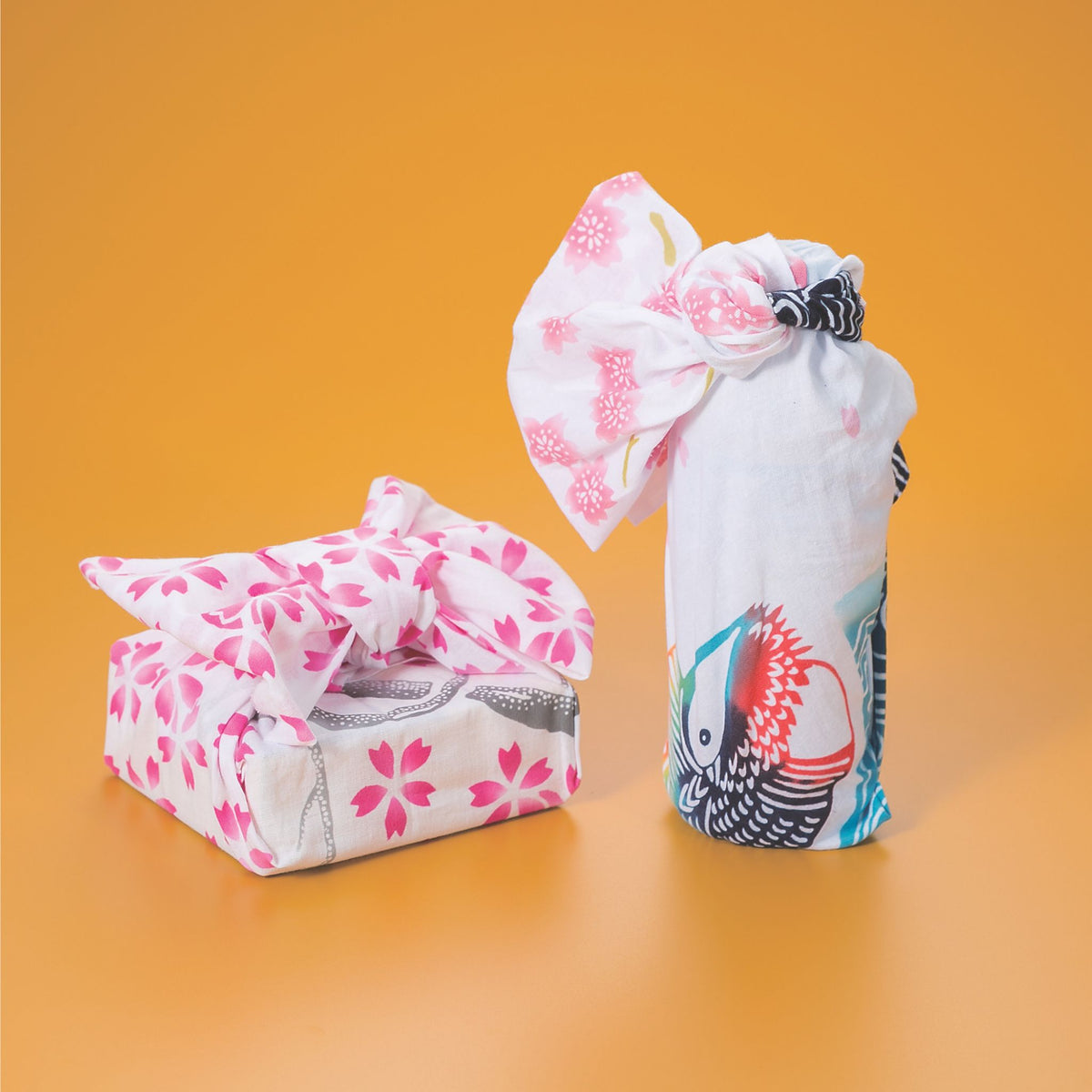 Two fabric gift wrap or pouches made from white lightweight fabric with pink floral prints and a koi fish design, displayed on a solid orange background.