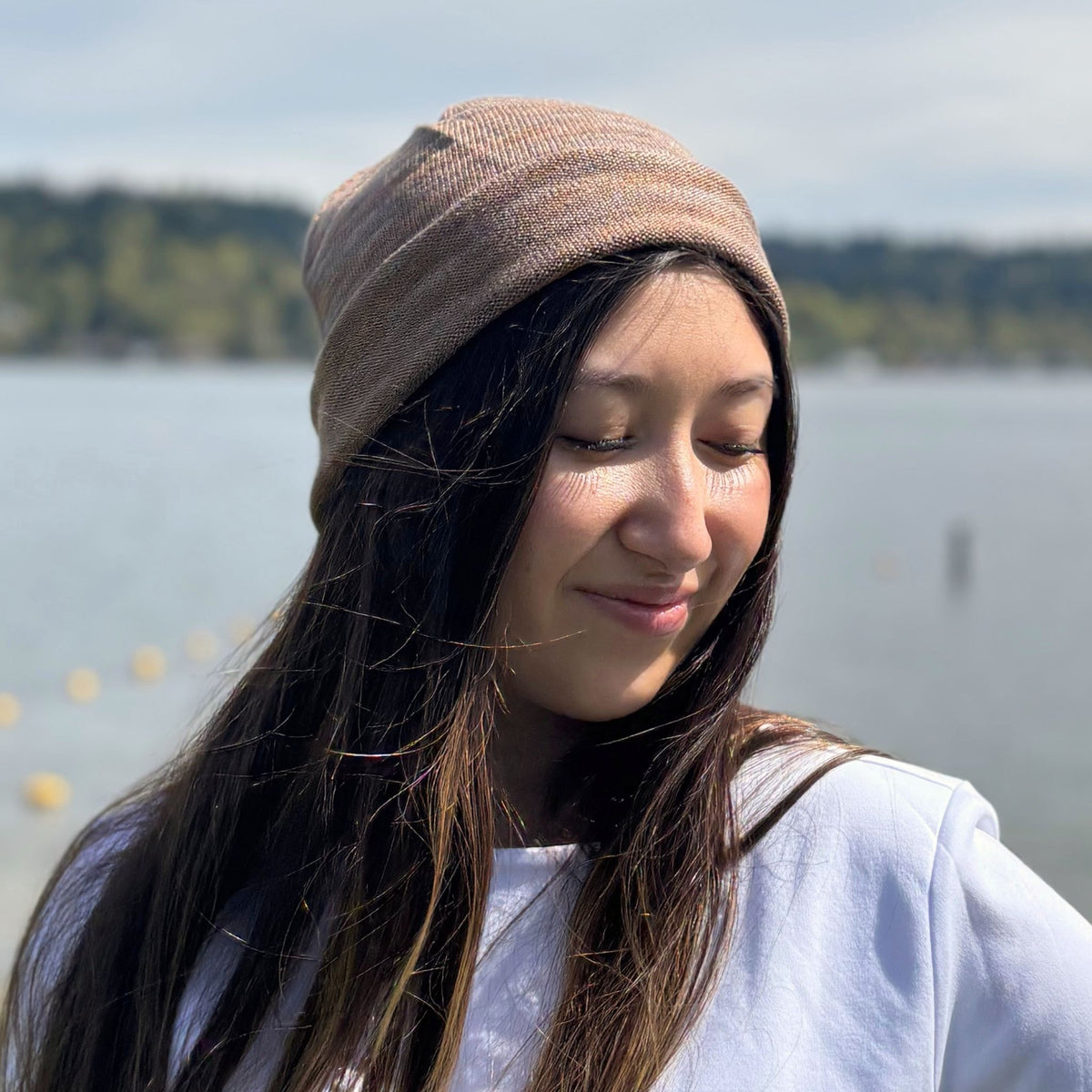 A woman wearing a brown silk beanie with a lake and trees in the background
