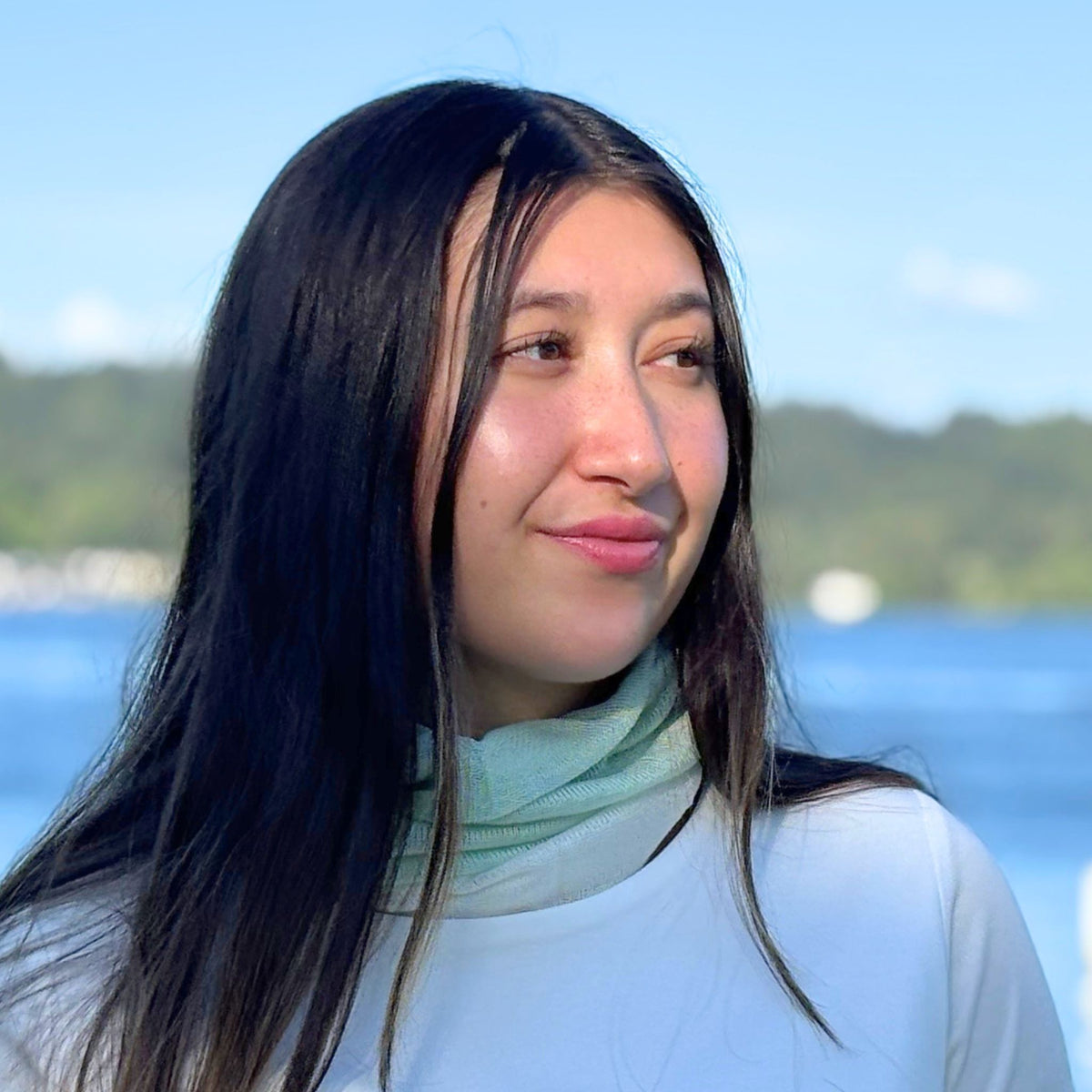 Woman with long dark hair and a light scarf standing in front of a lake with trees in the background.