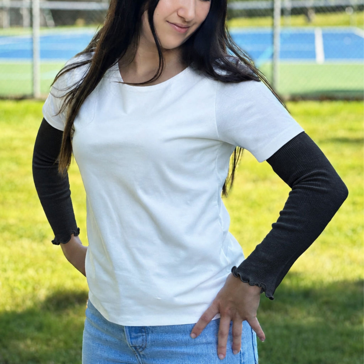 A woman standing outdoors on a grassy field with a tennis court in the background, wearing a white short-sleeve crewneck t-shirt with arm warmers, paired with light blue jeans.