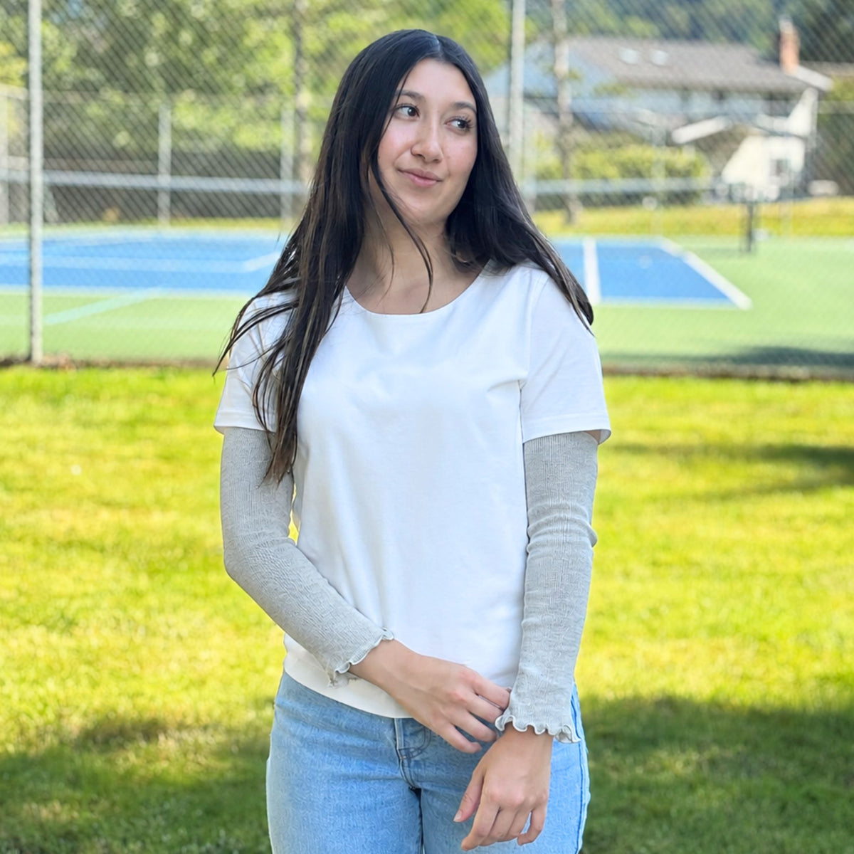 A woman standing outdoors on a grassy field with a tennis court in the background, wearing a white short-sleeve crewneck t-shirt with arm warmers, paired with light blue jeans.
