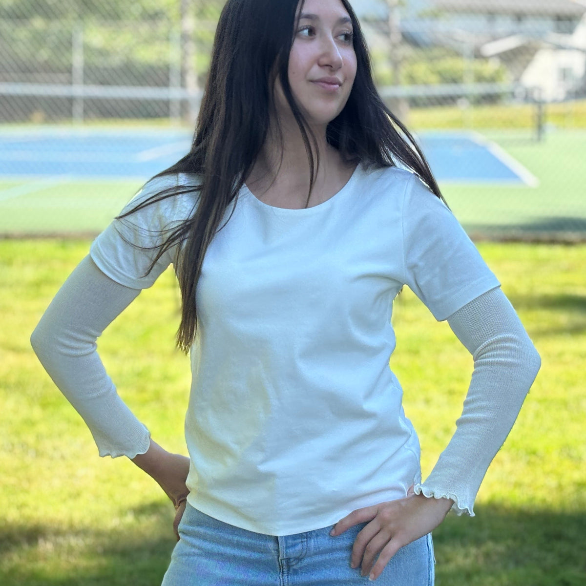 Woman wearing silk-blend-arm-cover and blue jeans standing on grass with a sports field in the background.
