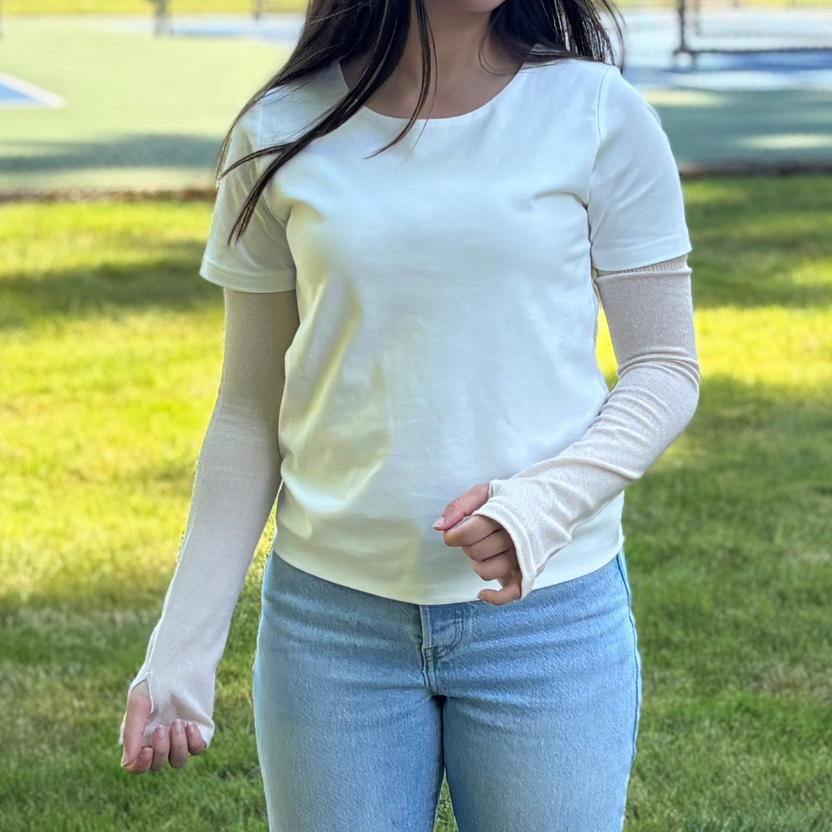 Woman standing on a grassy area wearing silk blend UV protection arm cover with tennis courts in the background