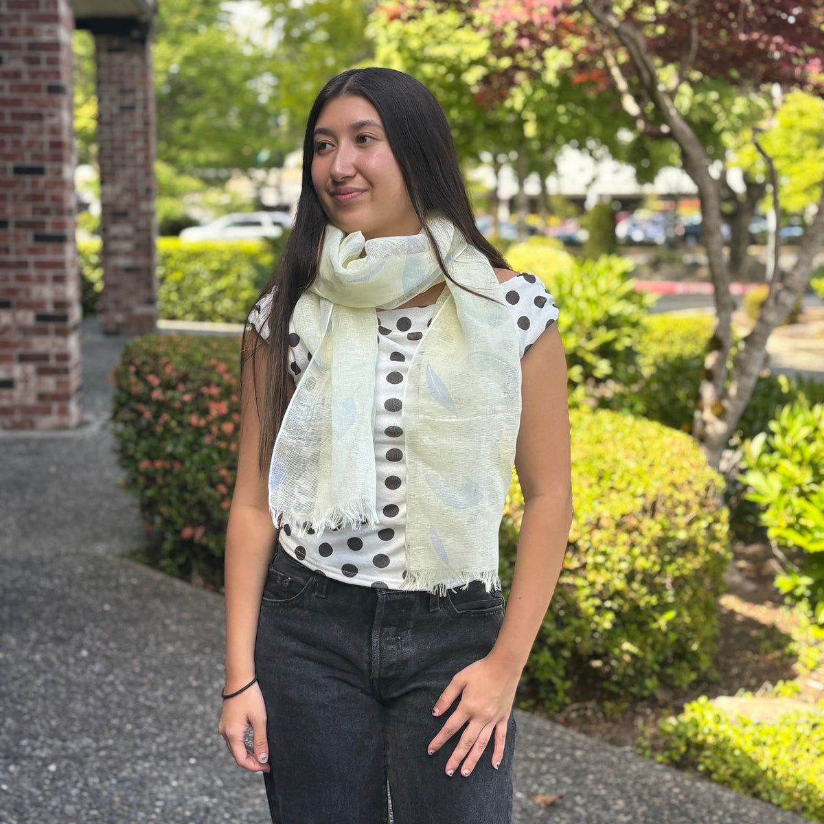 A woman standing outdoors on a paved walkway wearing a cream-colored scarf with black polka dots, a white sleeveless top with black polka dots, and dark gray jeans. Trees and a brick building are visible in the background.