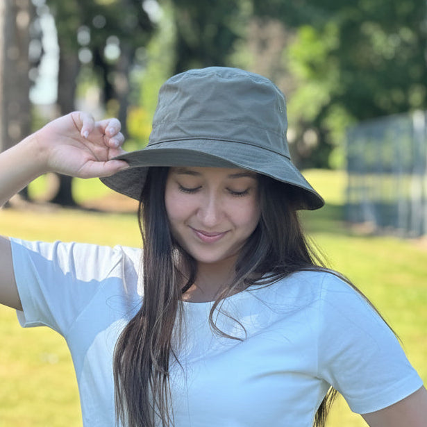 A woman wearing a light olive green wide-brim bucket hat and a white t-shirt, standing outdoors in a sunny grassy park with trees and a fence in the background.