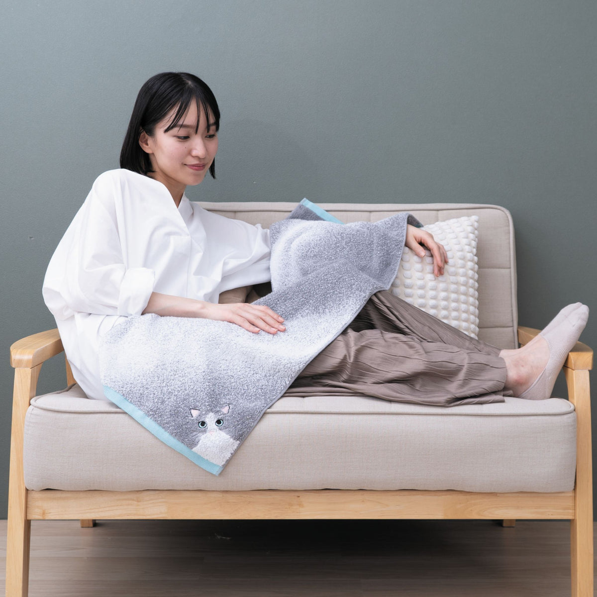 A woman sitting on a couch with a face towel draped over her, featuring a cat design.