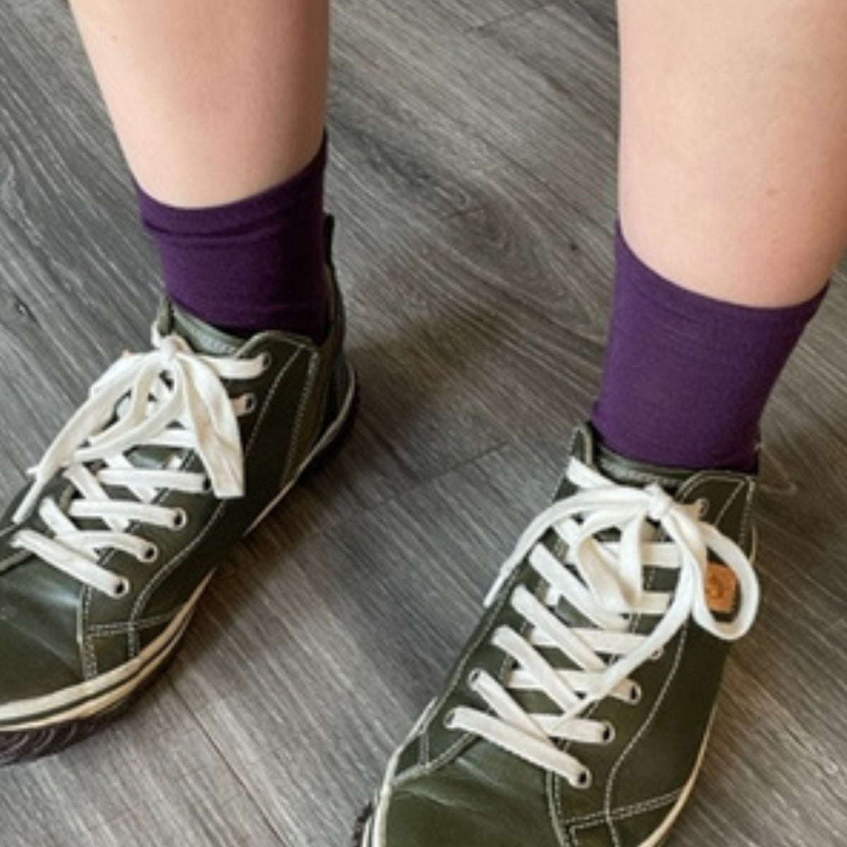 A close-up of a person's legs wearing purple high-top sneakers with white laces and purple crew socks, standing on a gray wood-look floor.