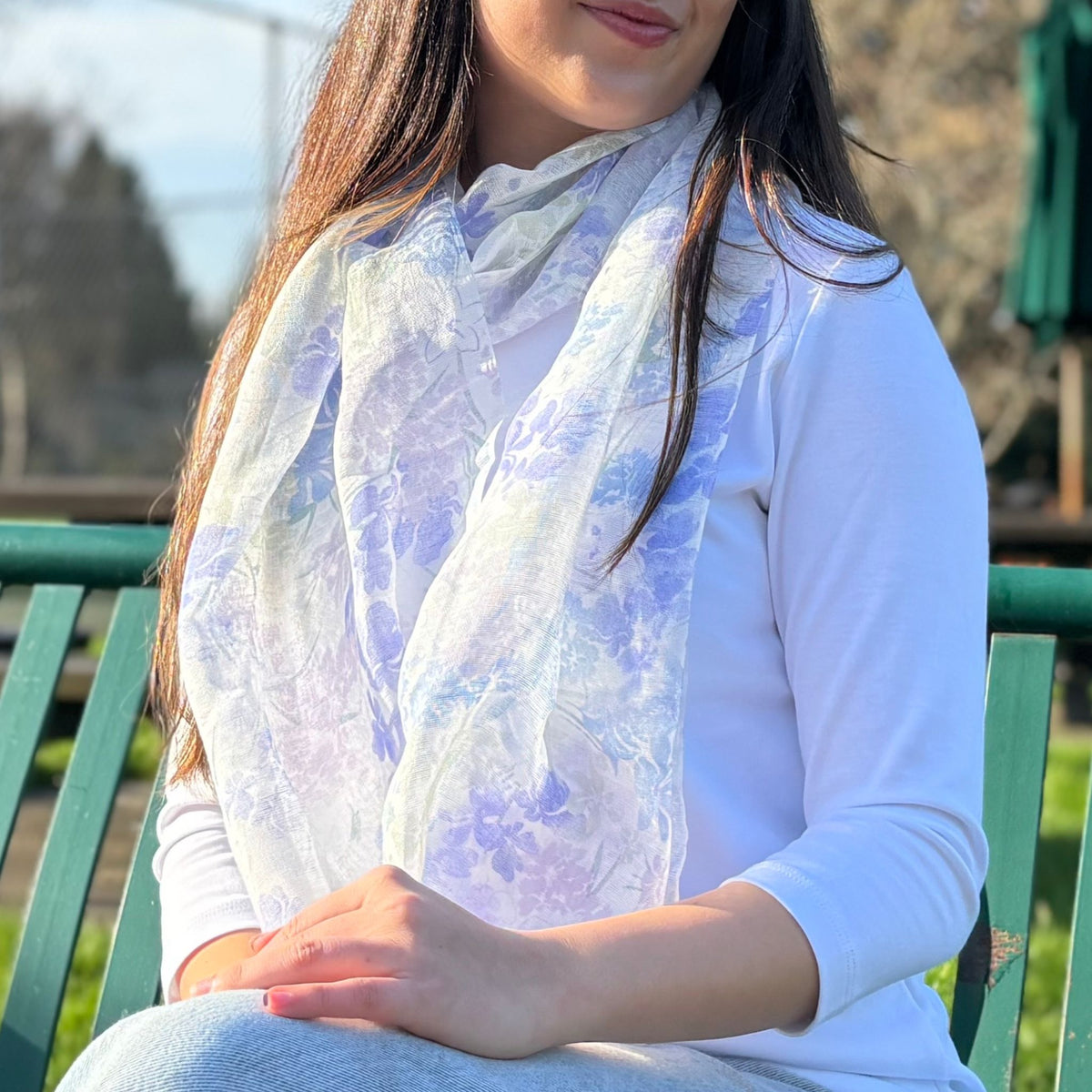 Person wearing a light purple floral scarf sitting on a green bench outdoors.