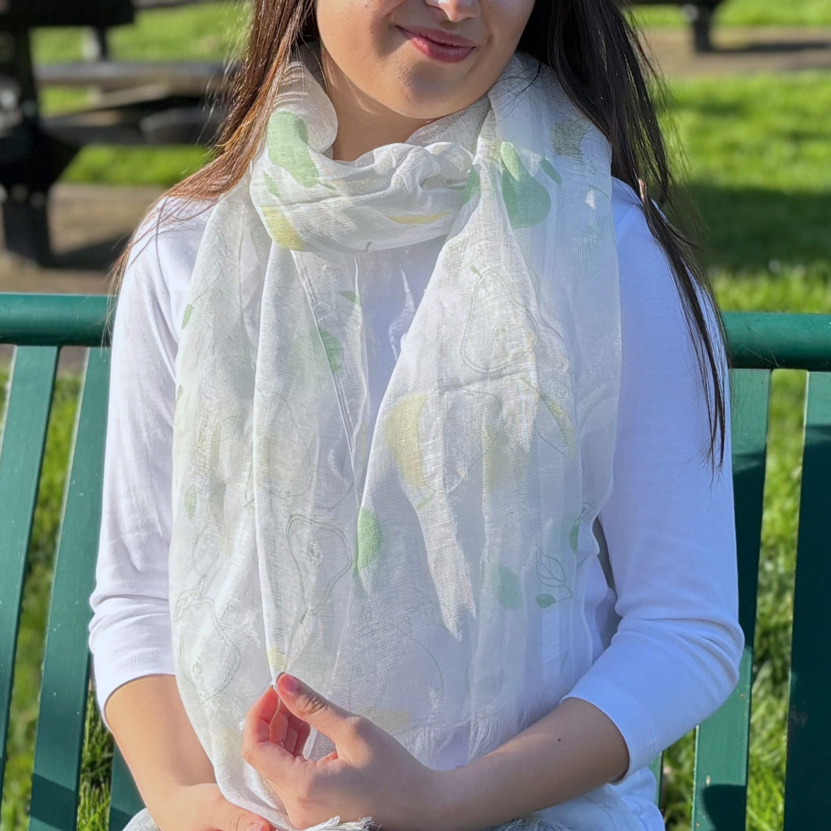 Person wearing a white scarf with green pear patterns, sitting on a green bench outdoors.