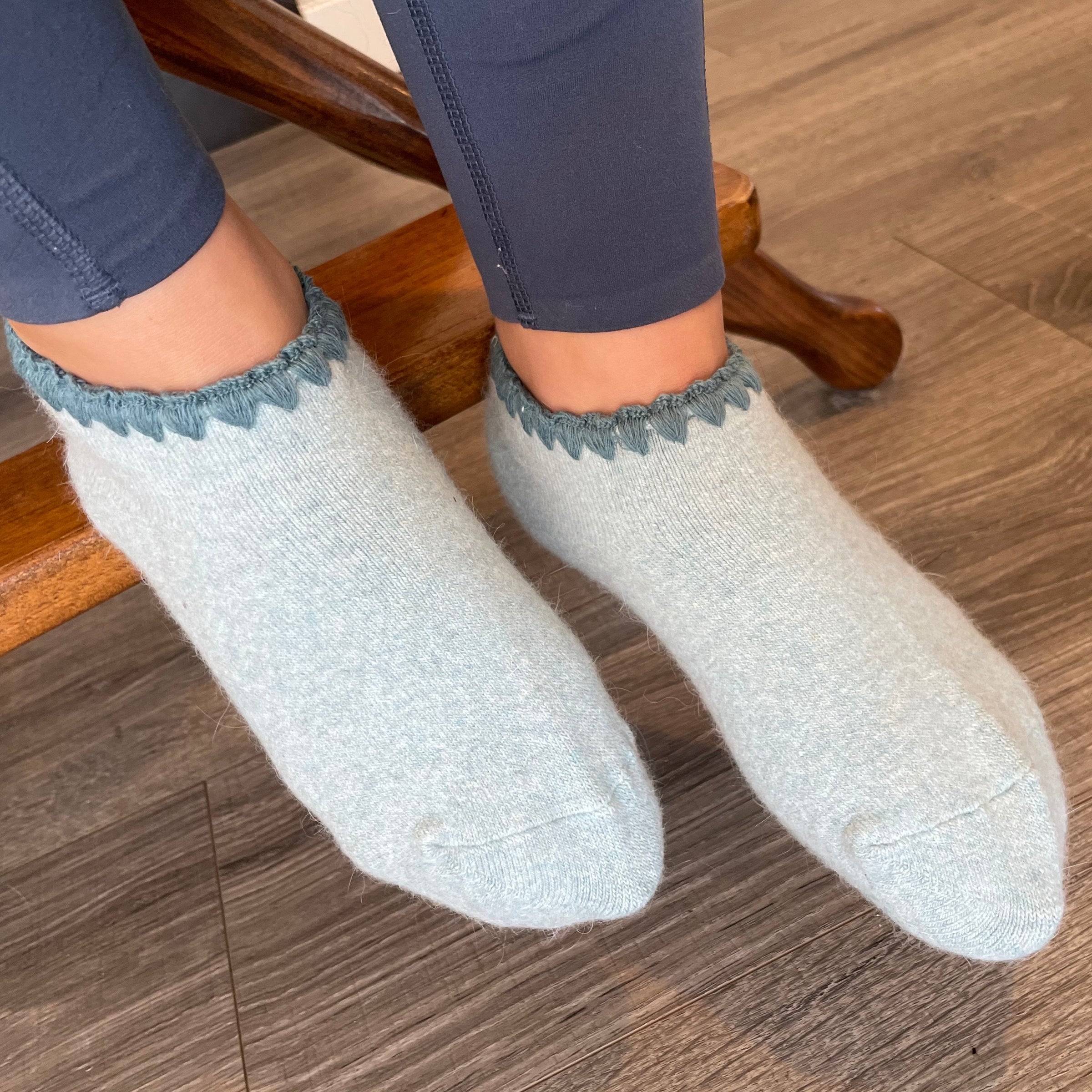 Angora Slipper Socks with anti-slip soles featuring transparent grip dots for extra safety. Each pair is handcrafted, incorporating traditional craftsmanship, and finished with cherrystone shell-shaped embroidery on the sock opening. Soft, cozy, and comfortable for ultimate foot pampering.